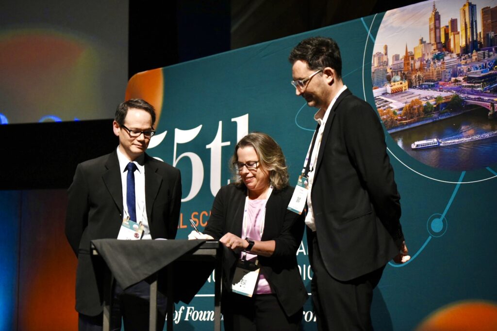3 people dressed professionally, on a stage, watch on as the woman in the middle signs a document on a lectern. They all look pleased.