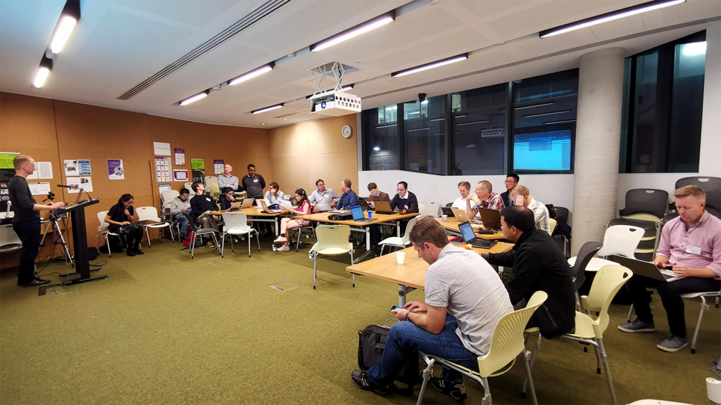 A seminar room in workshop configuration filled with people focussed on laptops and a presenter at a lectern